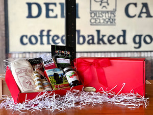 On the left, an open red gift box filled with treats and ingredients for a pancake breakfast. Pancake mix, maple syrup, ground coffee, energy bar, fruit preserves, and more. On the right, a closed red gift box tied with a box and a gold sticker. 